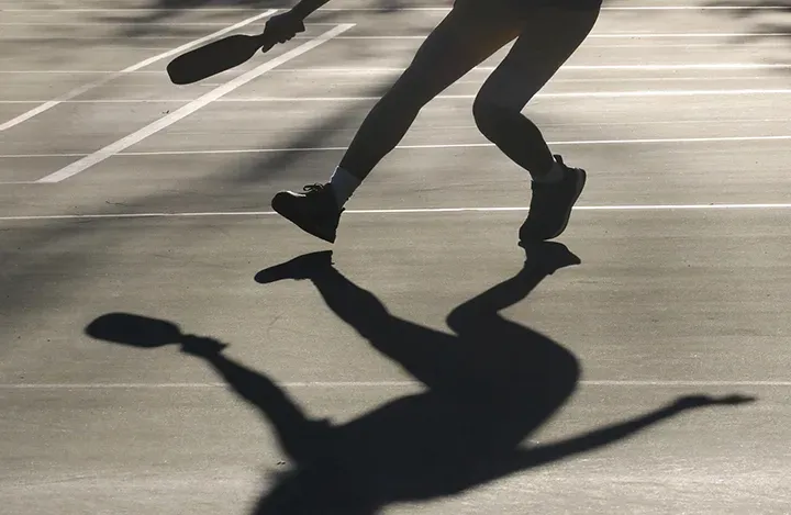 A player's legs and long shadow cast across the court in low afternoon sun, paddle raised