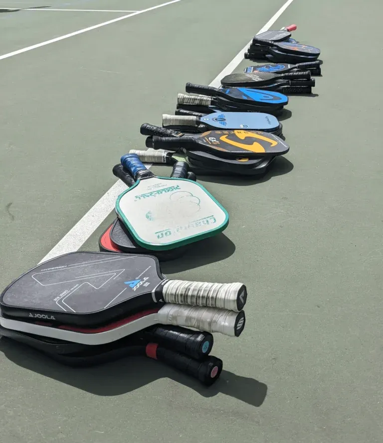 A long line of paddles stacked on the court surface along the baseline, the queue that decides who plays next